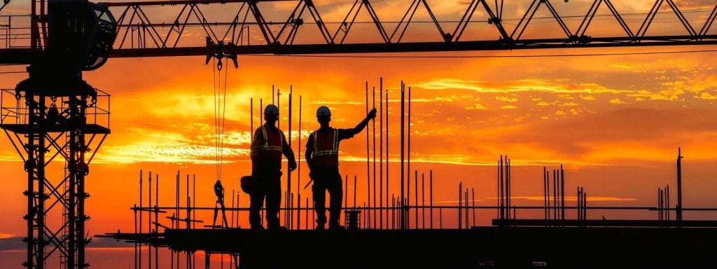 Construction workers silhouetted against a sunset, overseeing crane operations and lift planning on a building site in Boston.