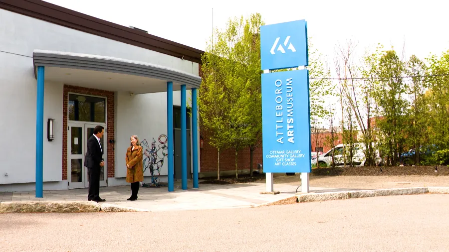 Attleboro Arts Museum entrance with two people conversing, featuring a blue sign displaying "ATTLEBORO ARTS MUSEUM" and surrounding greenery, highlighting the cultural landmark in Southern Massachusetts.