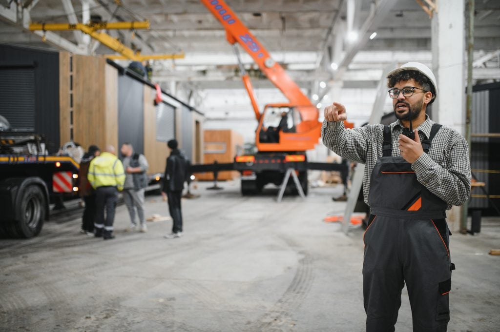 Arab engineer supervising crane operations in a construction site, discussing lift planning with a team, featuring modular units and heavy equipment in the background.