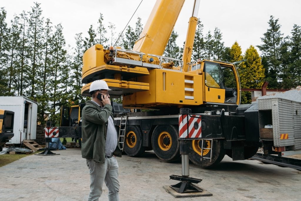 Southern Massachusetts: A construction worker in a hard hat talks on his phone in front of a large, yellow crane at a construction site.