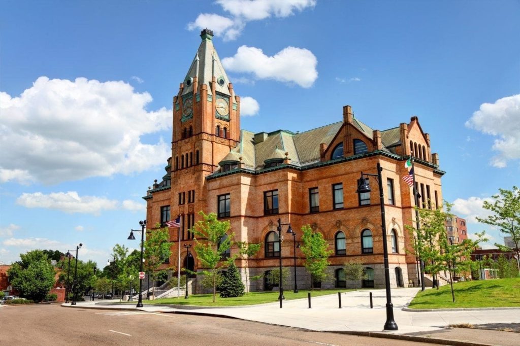 Brockton City Hall, a historic brick building with a clock tower, surrounded by green trees and street lamps, showcasing the local architecture and community atmosphere in Brockton, MA.