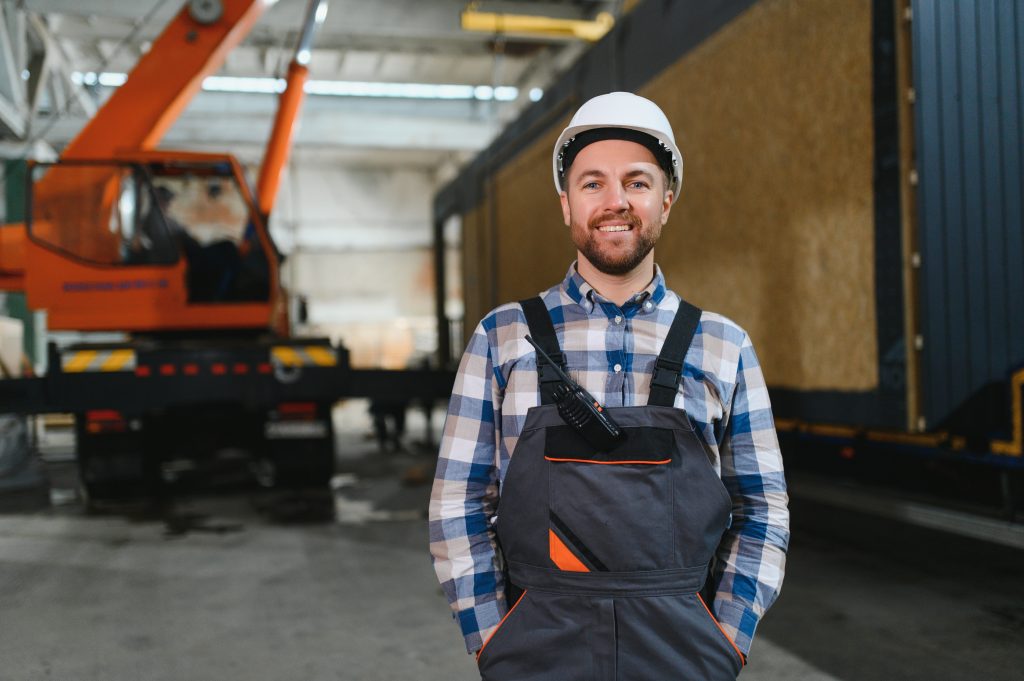 Smiling male crane operator in safety gear, standing in front of a crane in a warehouse, emphasizing expertise in crane rental services for safe and efficient lifting in Raynham, MA.