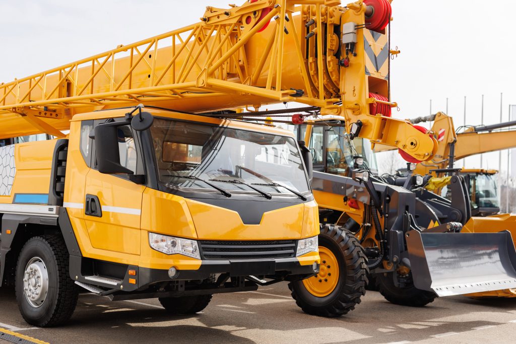 Self-propelled crane with yellow boom and heavy machinery in construction yard, representing MacDonald Crane's rental services for efficient lifting and rigging in Bridgewater, MA.