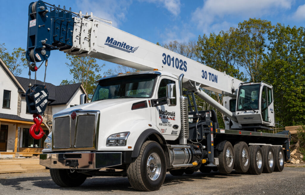 30-ton boom truck from MacDonald Crane parked at a residential construction site, featuring a hydraulic boom and lifting hook, surrounded by trees and a partially built house.